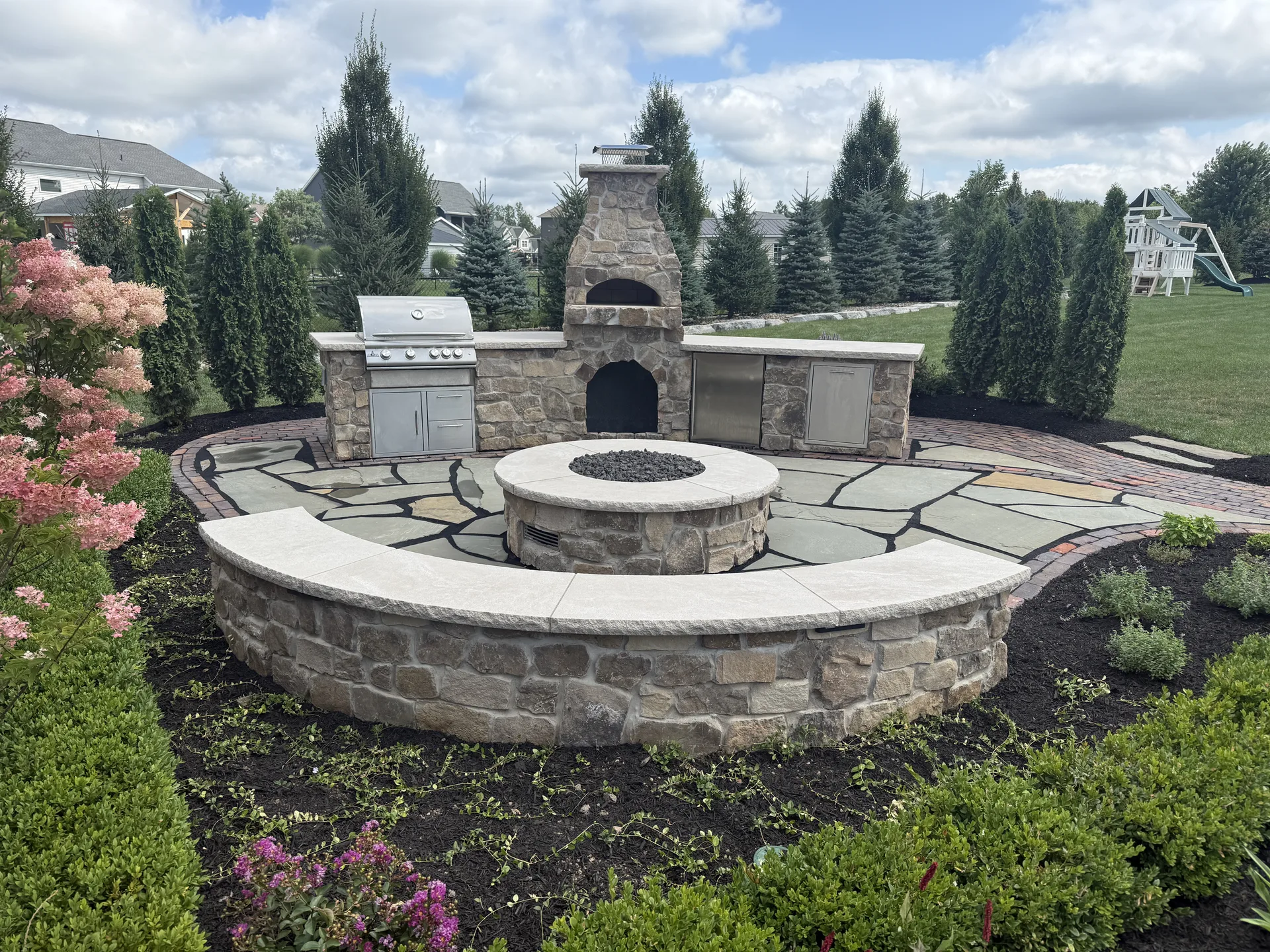 Stone outdoor kitchen, fireplace, fire pit, and curved seating wall surrounded by planting
